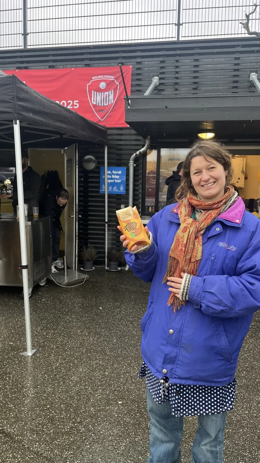 A smiling supporter in a blue coat at Boldklubben Union, holding a branded Sloppy Jo's Hotwrap carton with a hot Flatwrap inside, the Union banner behind.
