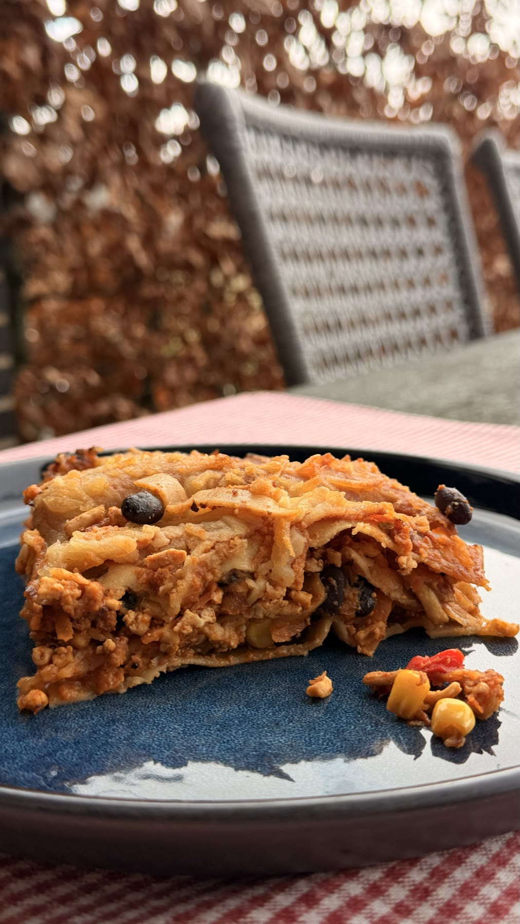 A tall profile of a Mexican lasagne portion plated outdoors, black beans and corn visible in the layers, a bright red-and-white checked napkin peeking at the edge